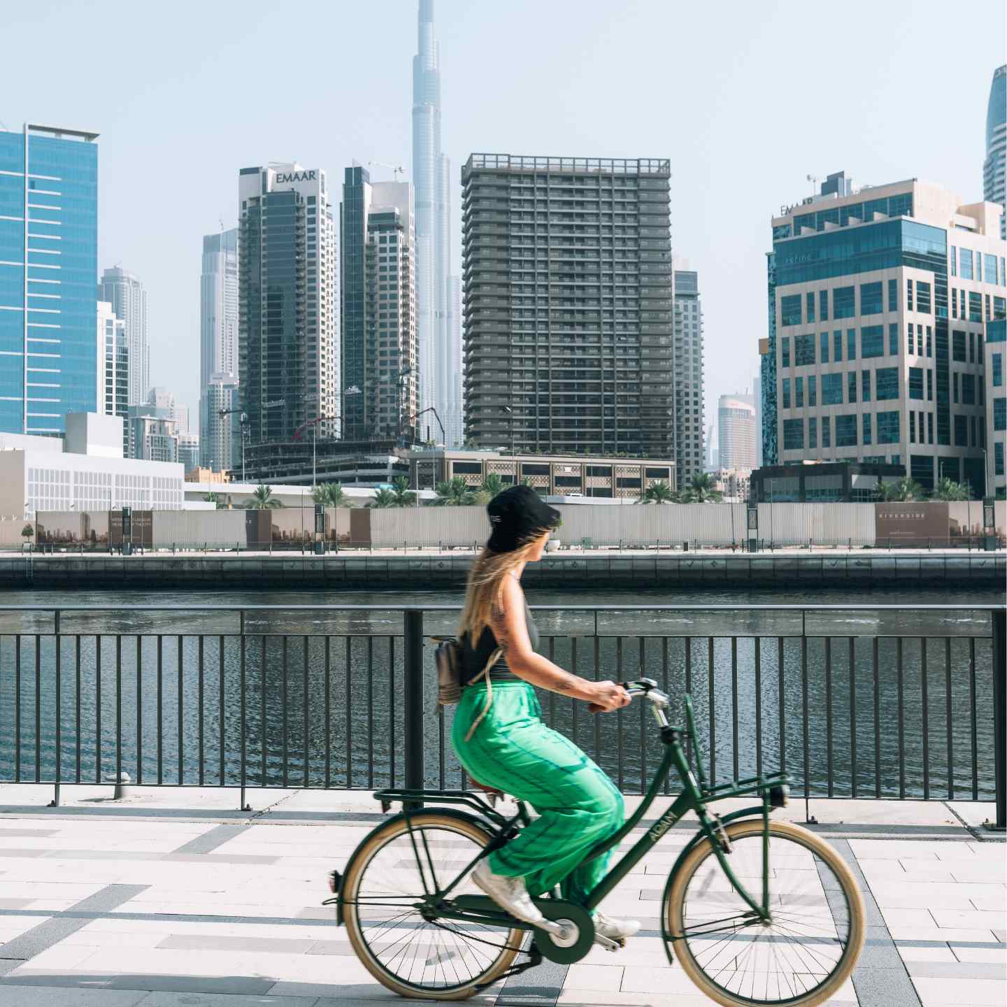 Une femme en jupe verte faisant du vélo le long du front de mer de Dubaï, avec un canal et des gratte-ciels en arrière-plan