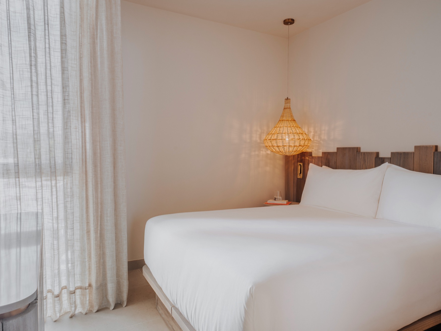 A double bed with bedside table and ceiling lamp on the right, with a window on the left covered by sheer curtains