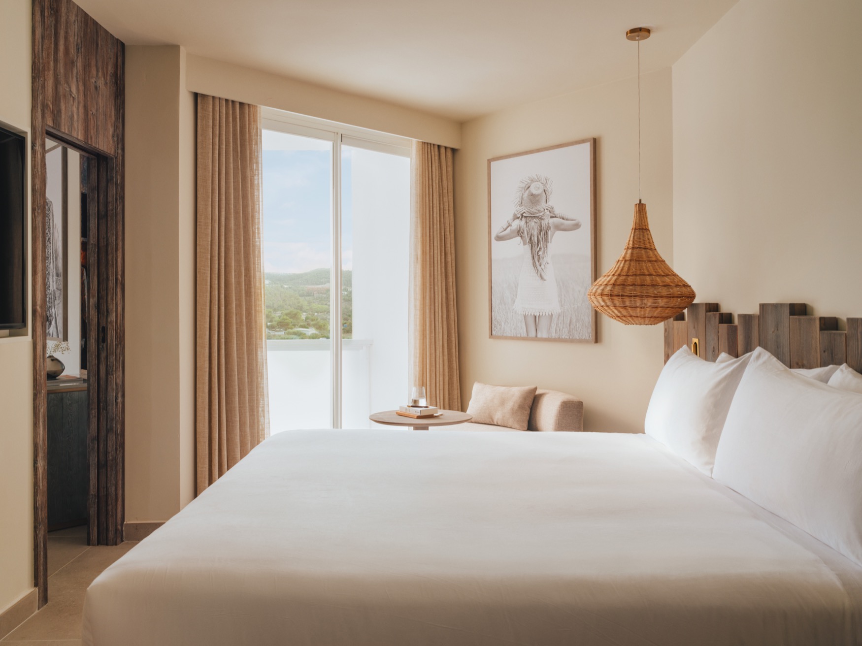 A double bed in the forefront with bedside table and ceiling lamp, with a balcony view at the background