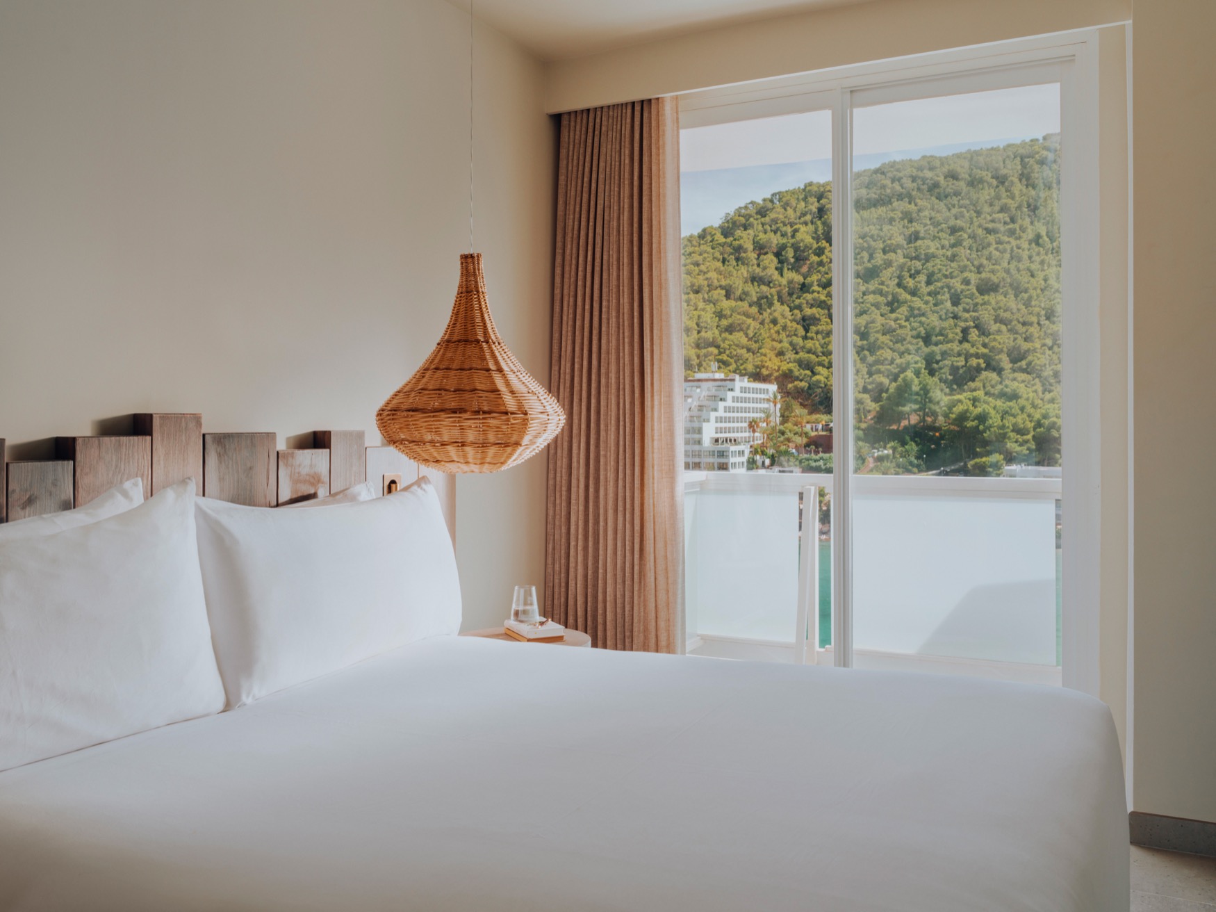 A double bed in the forefront with bedside table and ceiling lamp, with a balcony view at the background showing green hills