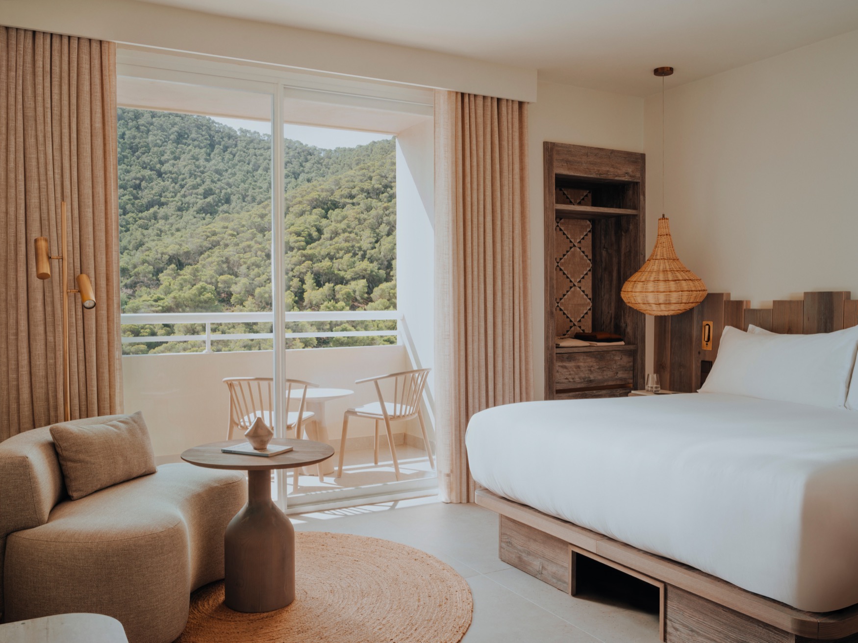 A small couch with coffee table on the left, a double bed with lamp and wardrobe space on the right, showing a balcony at the centre of the image with 2 chairs and a view of green hills