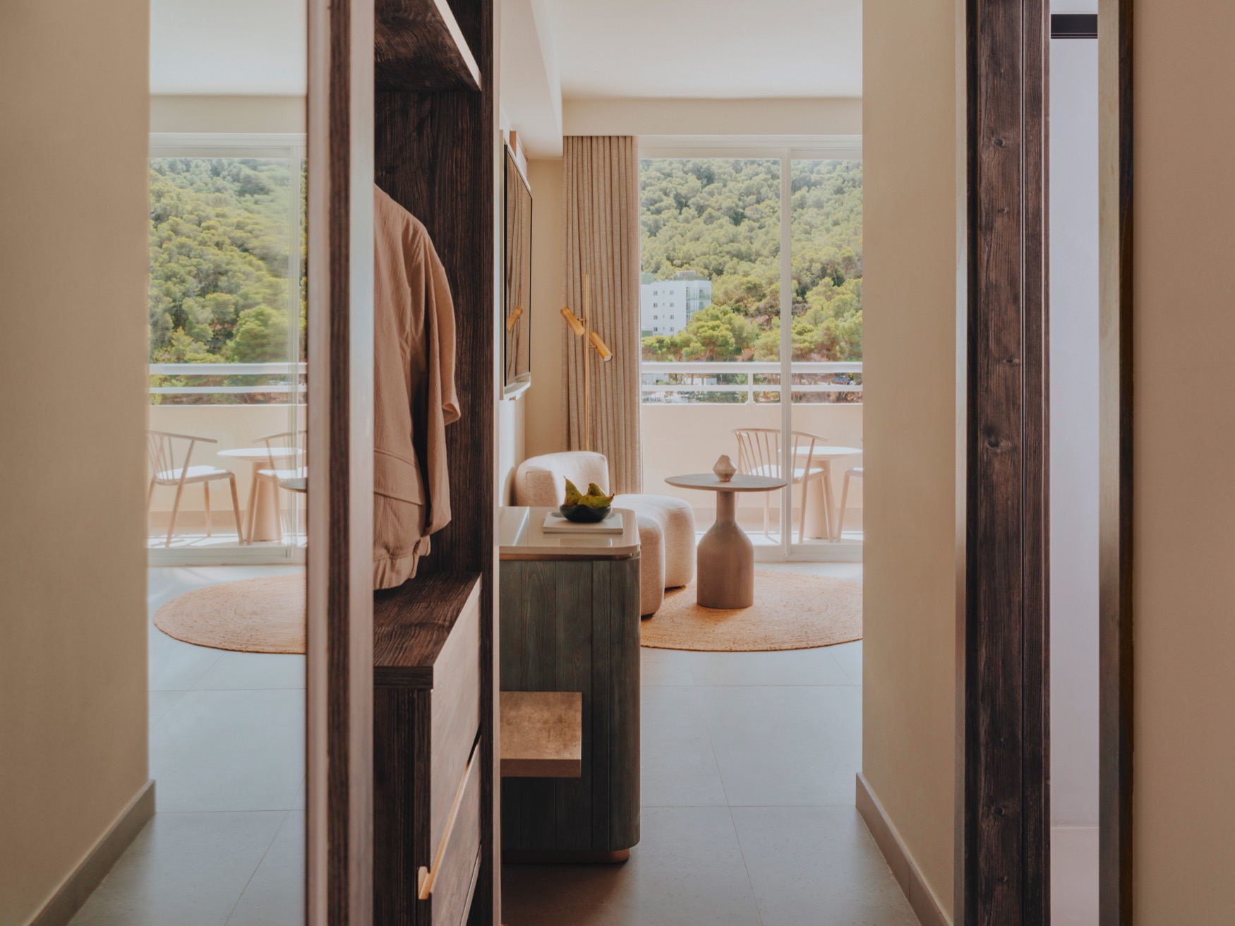 Vue d'un couloir avec penderie intégrée sur la gauche avec des vêtements suspendus, une porte de salle de bain sur la droite, et vue sur une coiffeuse, un fauteuil, une table basse et un balcon au centre de l'image