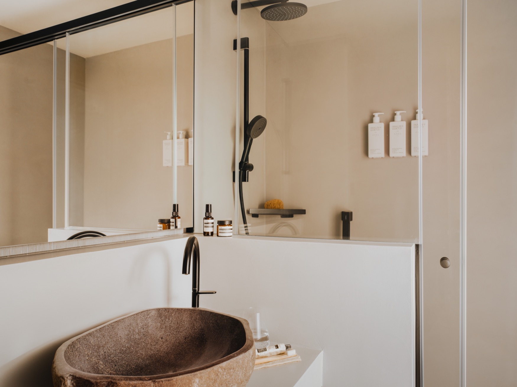 A bathroom with a sink and walled mirror at the forefront, with an open shower in the background