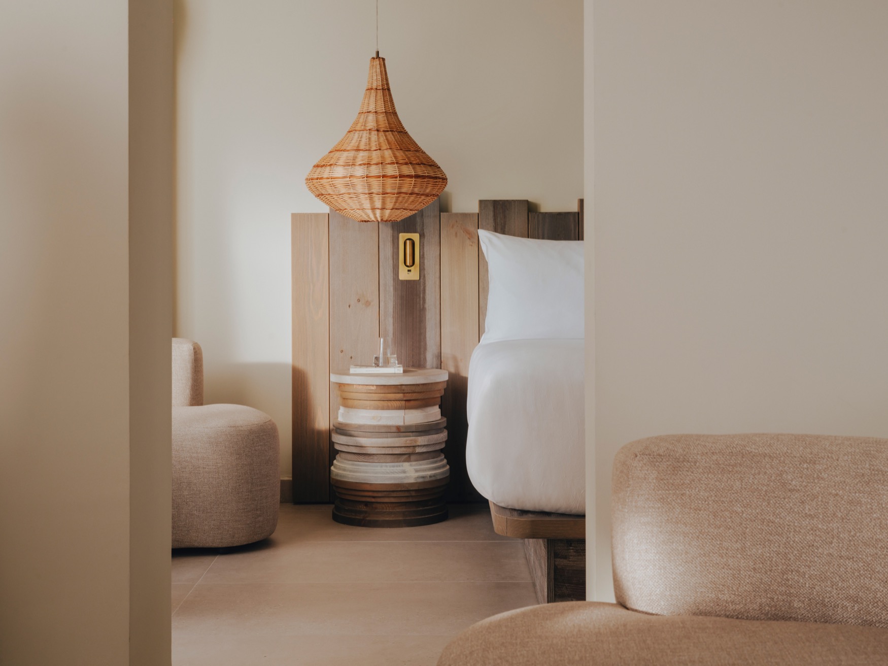 A soft armchair in the forefront, followed by a partial display of double bed, bedside table, a ceiling lamp and another armchair in the background