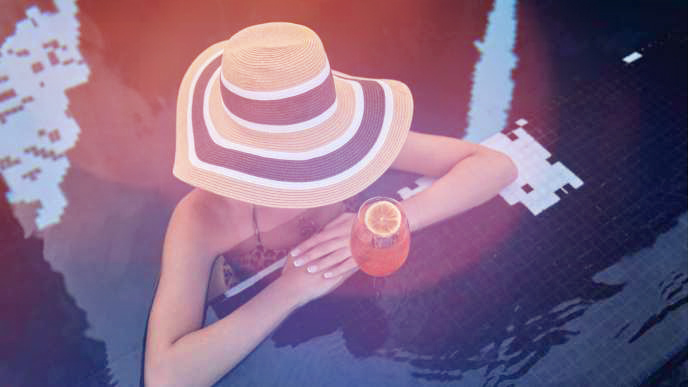 Femme portant un bob, les bras sur le rebord de la piscine avec un cocktail orange à côté d'elle