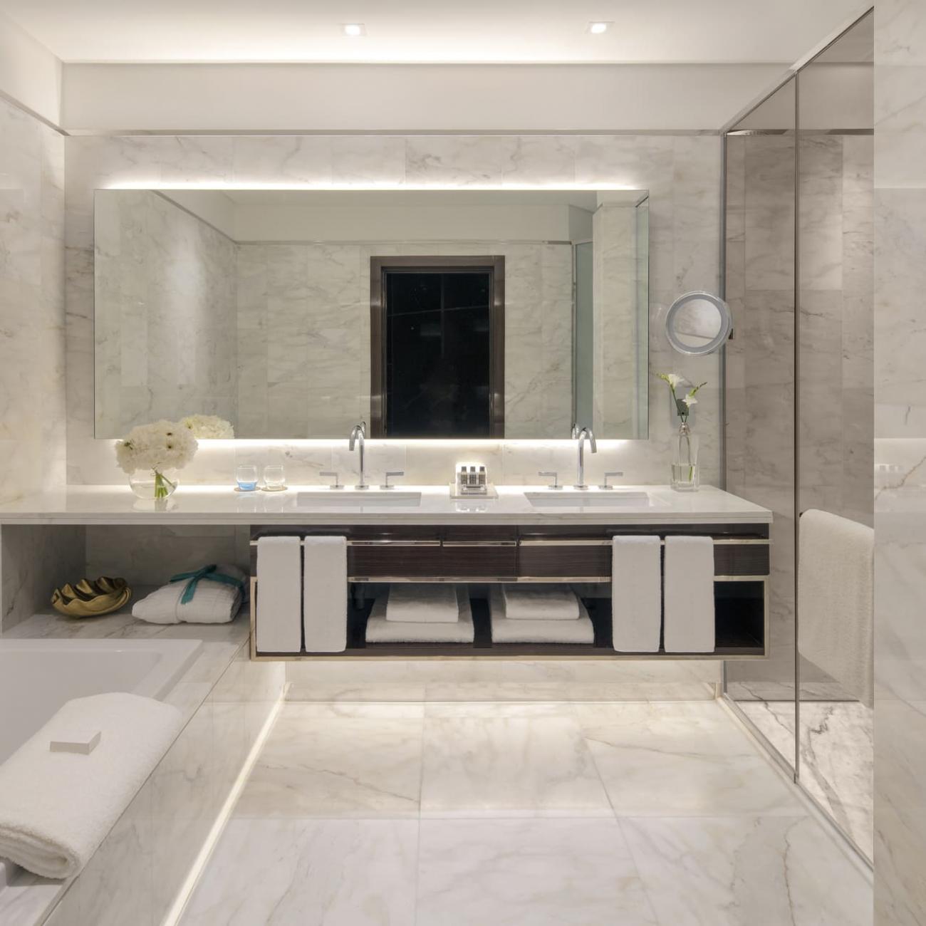 A large marbled bathroom with 2 sinks and drawers in the background, with a bathtub on the left and full mirrored wall on the right.