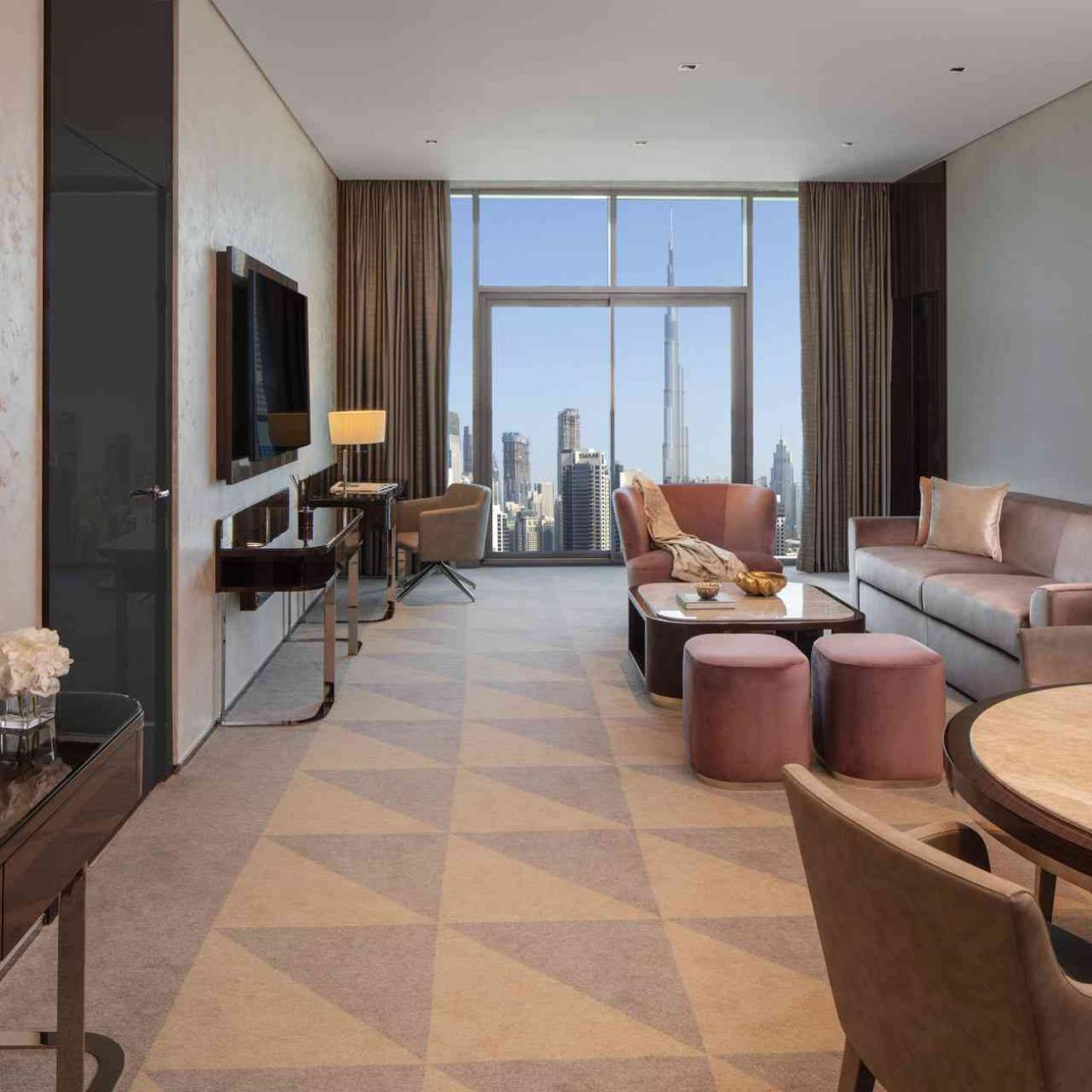 A living room displaying a part of round dining table and chairs in the foreground, with a couch, armchair, 2 ottomans and a coffee table in the middle of the room opposite from the wall hanged TV, with the background of a top to bottom window showing views of Dubai and Burj Khalifa building