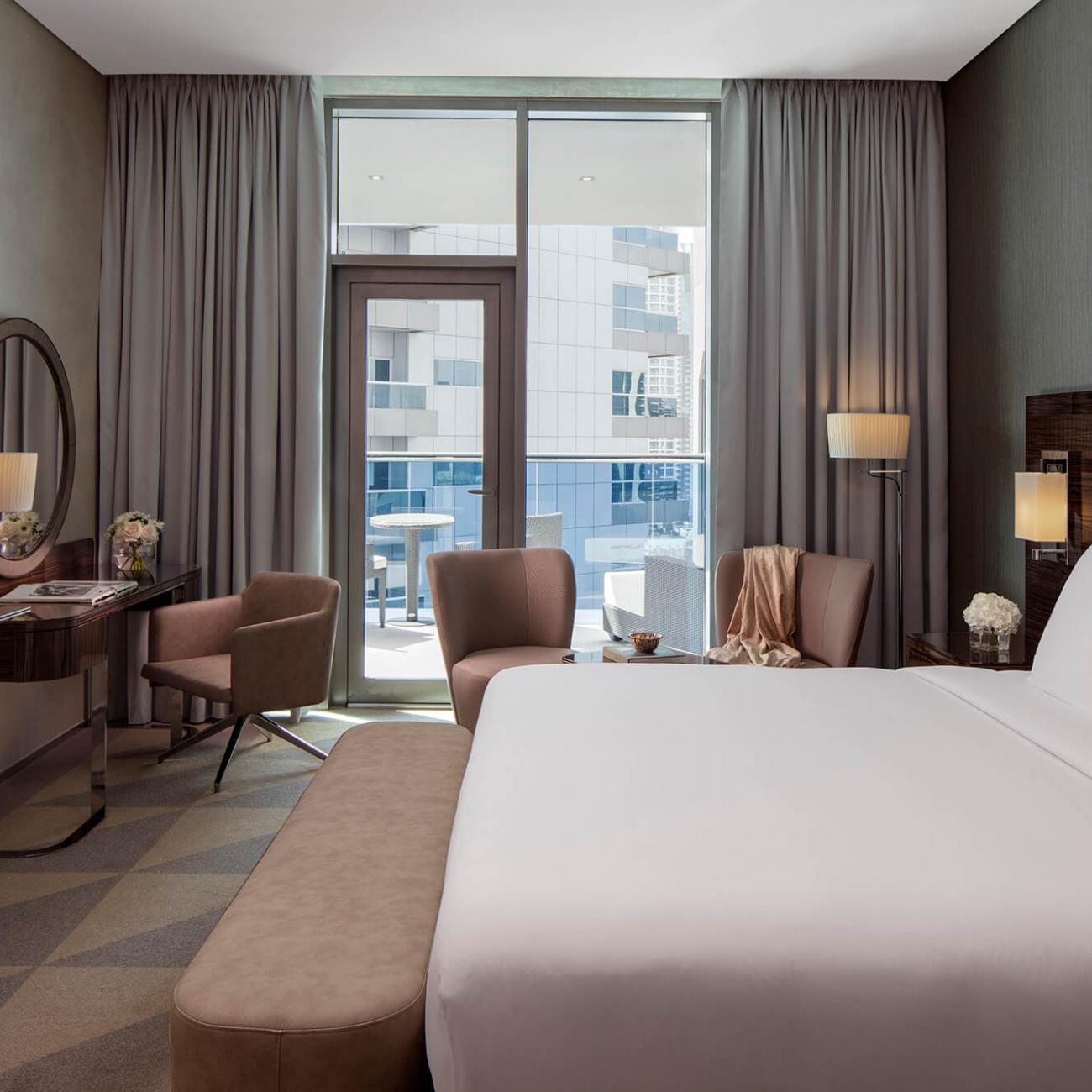 A king size bed with a bedroom bench in the foreground, with a dresser table and mirror on the left, 2 occasional chairs with coffee table and floor lamp, finished by a balcony view in the background