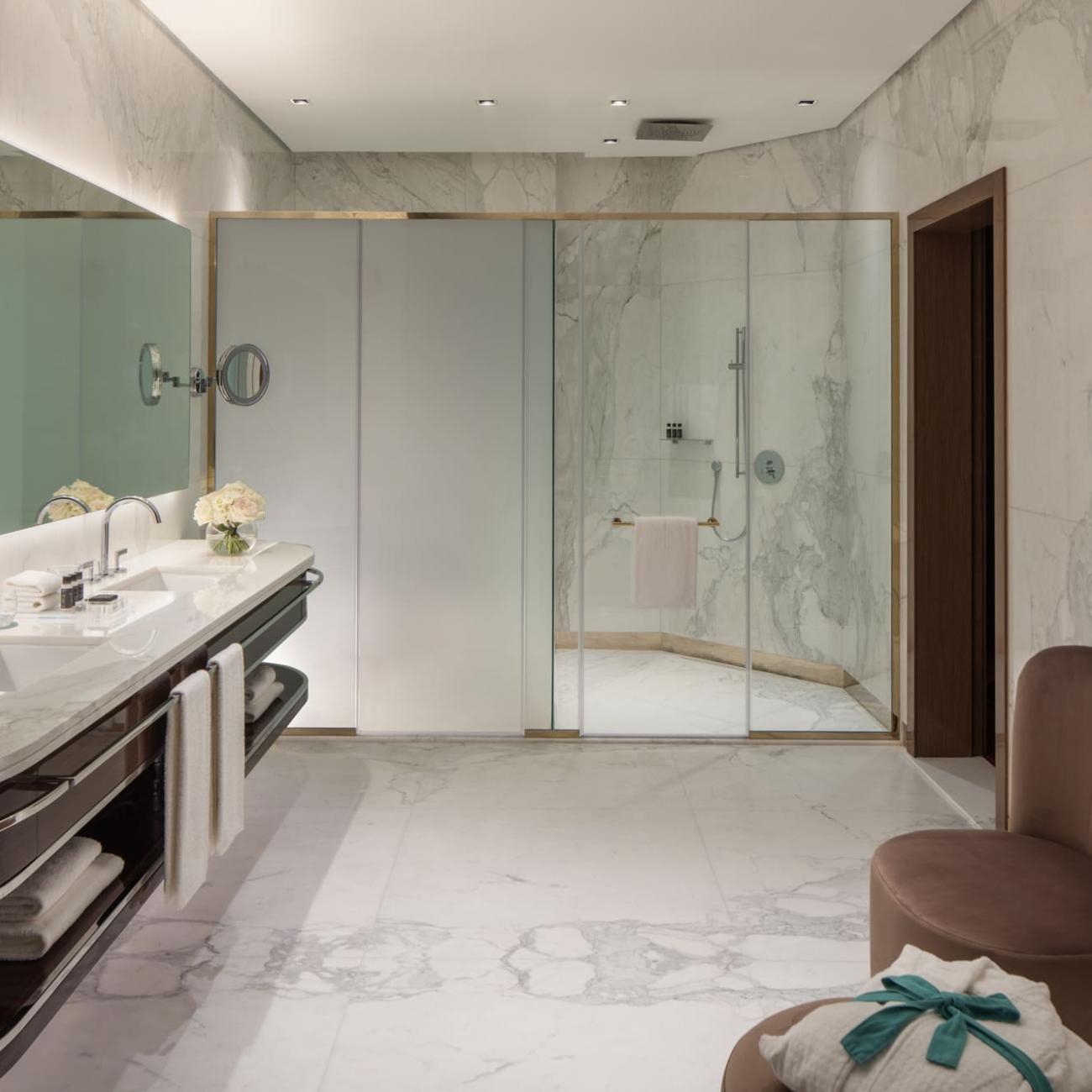 Large marbled bathroom with two sinks on the left, large walk in shower in the background, and entrance door and an armchair with a side table on the right