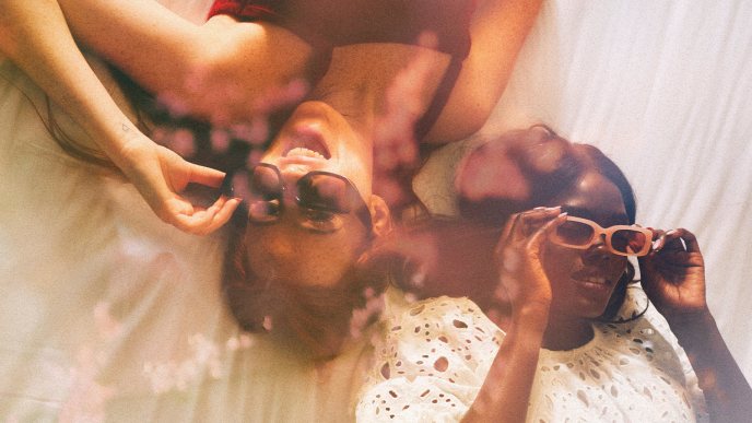 Two girls lying on a white bed putting on sunglasses