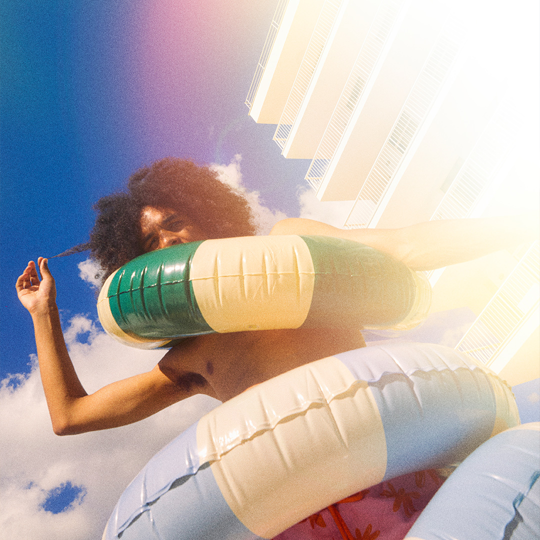 Man with curly long hair wearing two pool float rings around his body with blue sky in the background