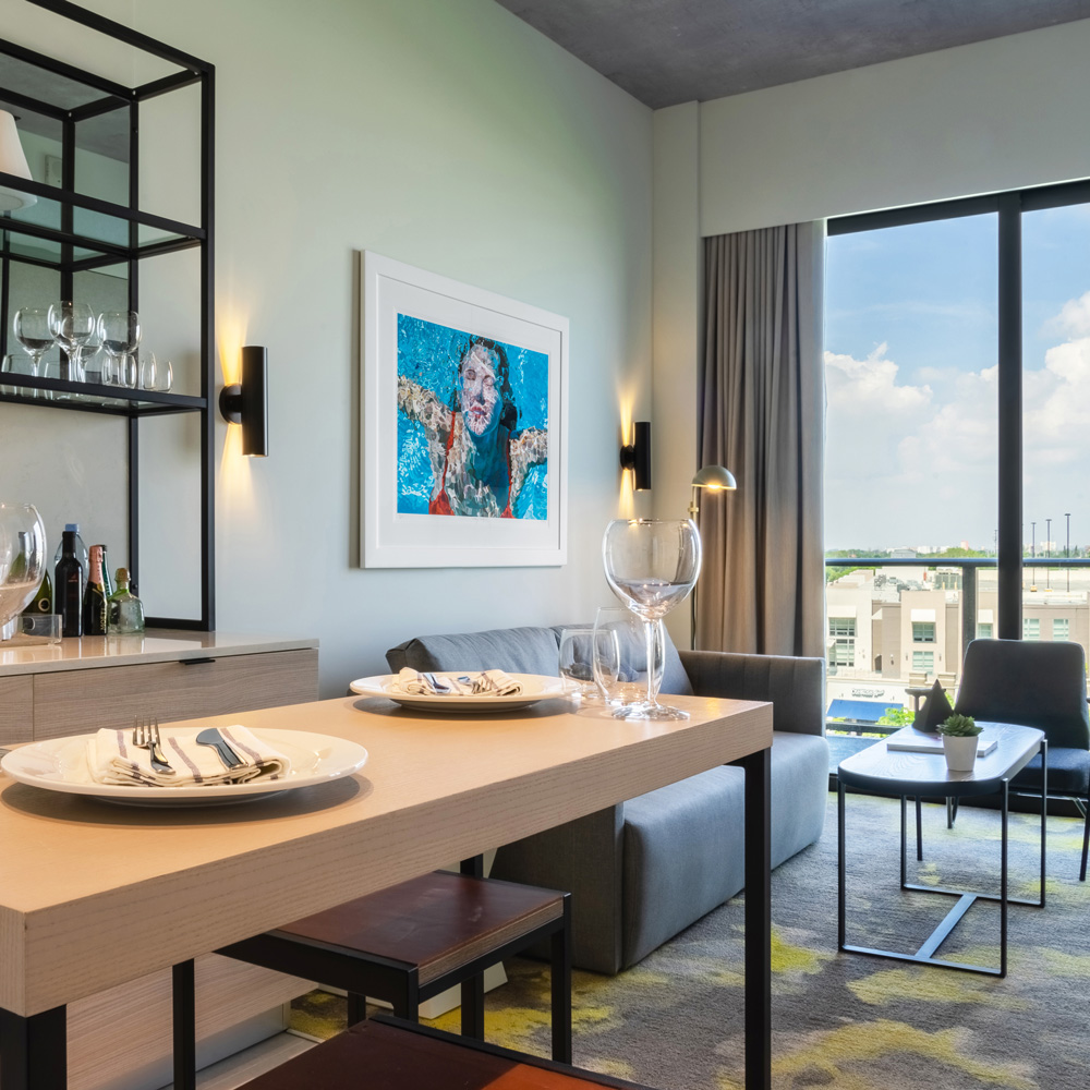 Dining table set for two in a living area for a hotel suite, with sofa and coffee table in the background