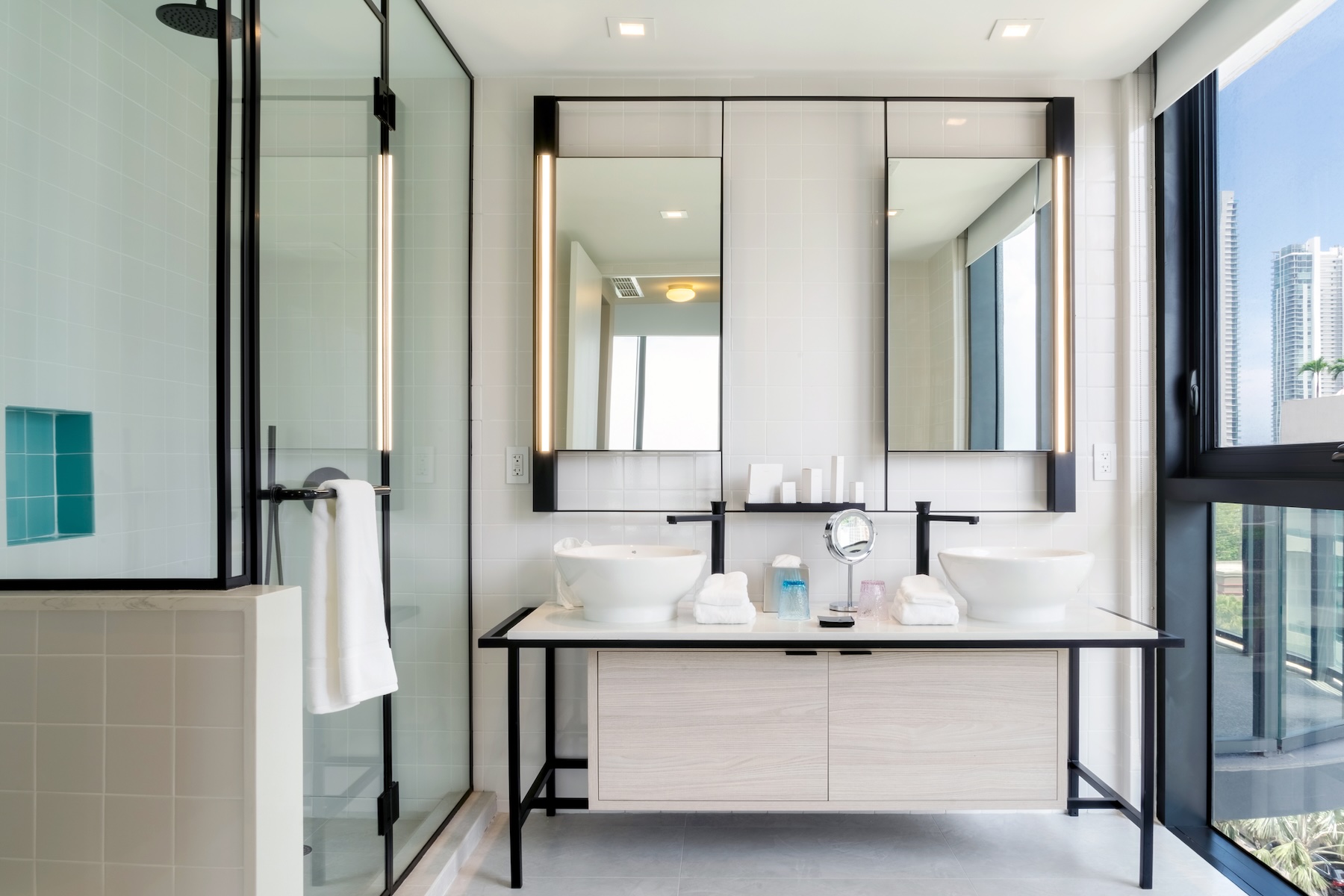 Salle de bain moderne avec double vasque au centre de l'image, douche à l'italienne vitrée sur la gauche, et grande baie vitrée avec vue sur la ville