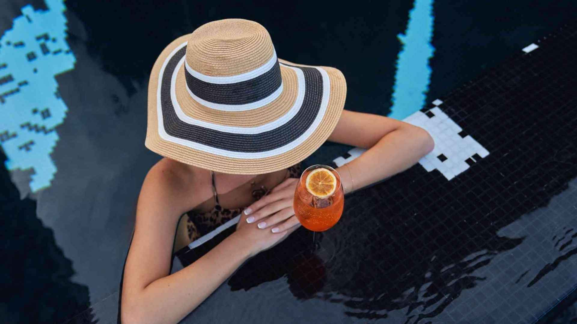 Femme au chapeau rayé accoudée au bord de la piscine avec un verre de spritz orange