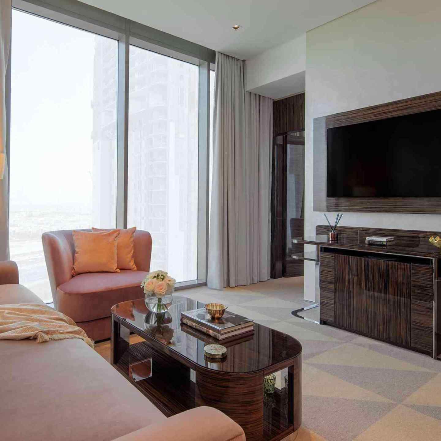 Pink sofa and pink lounge chair with dark brown coffee table facing a large tv on a wall and floor to ceiling windows in the background