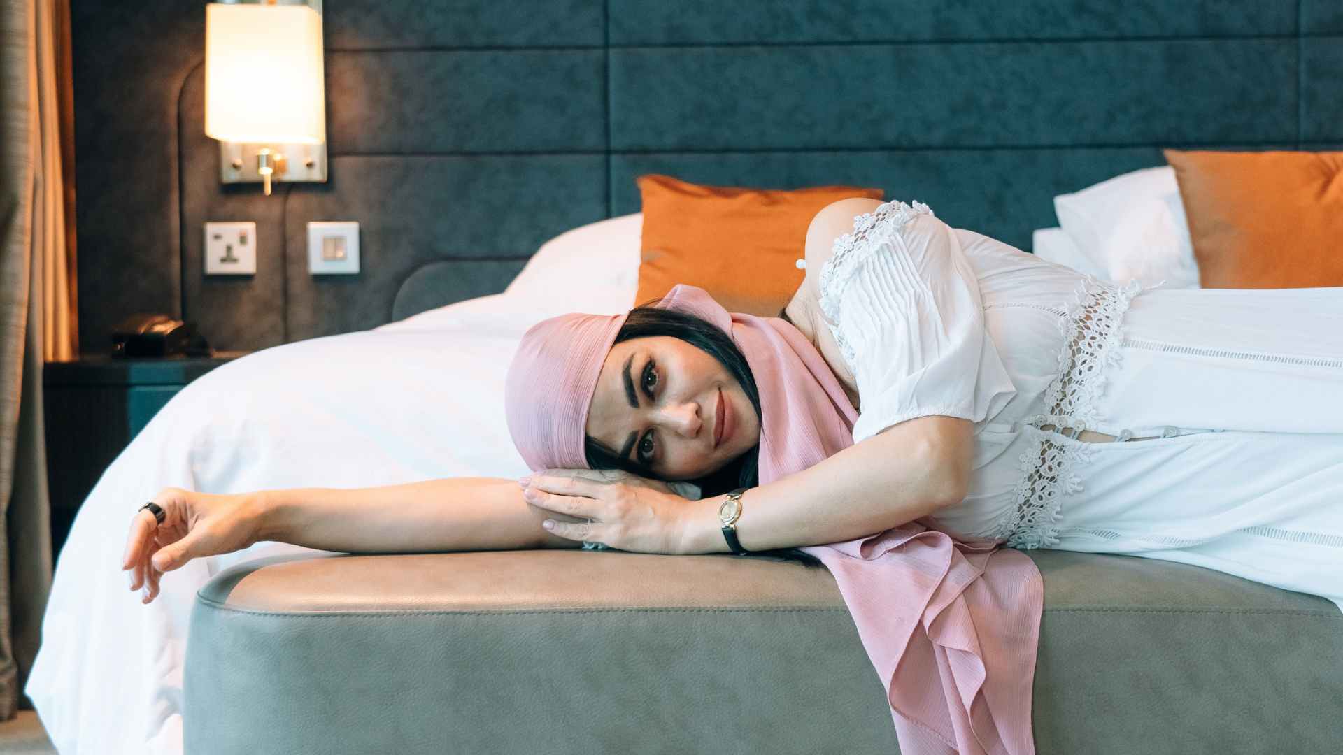 Woman in pink head wrap and white dress lays on a gray bench at the end of a bed