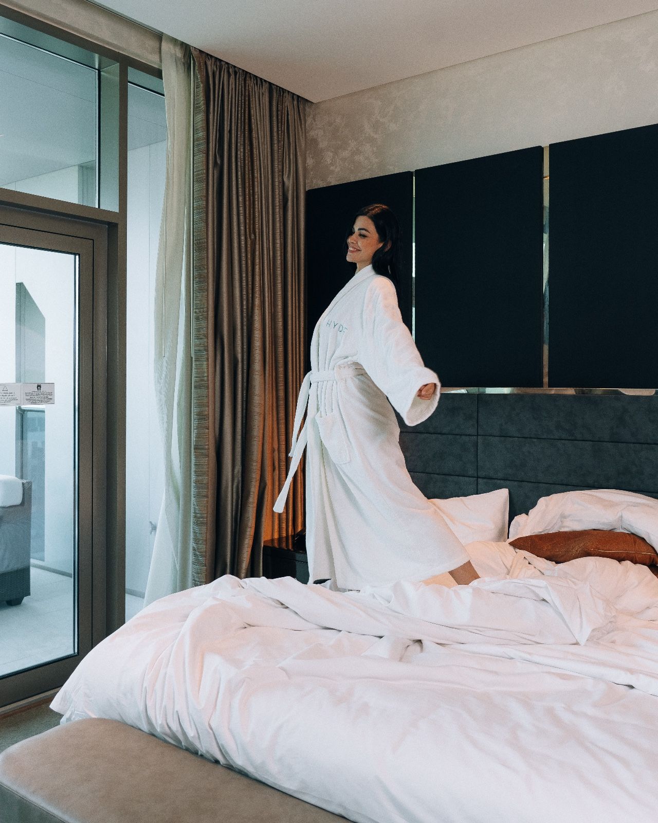 Woman in white robe stands on a white bed with grey headboard and gold curtains in background