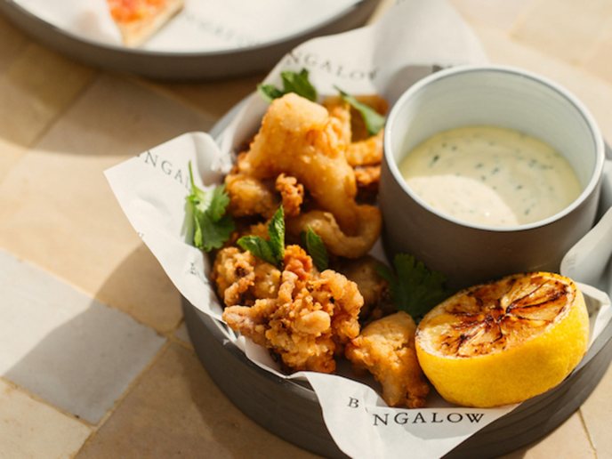 Calamari in a bowl with dipping sauce and lemon wedge with pool and pan con tomate in the background