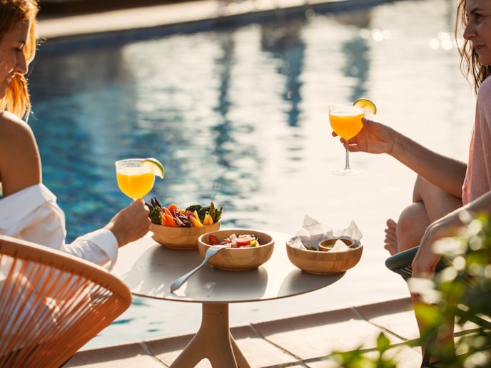 Friends with orange cocktails in hands and small table of food in the background and pool