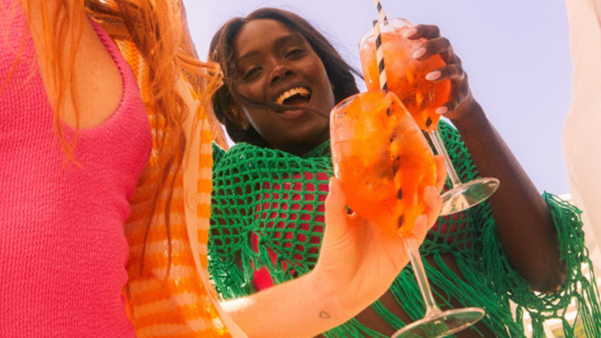 Woman in green fishnet cover up smiles while holding a drink, woman's body next to her in pink bathing suit also holding a drink