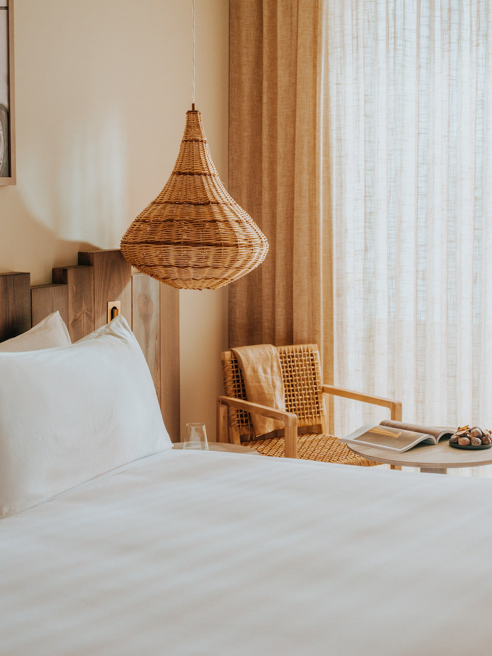 Close up of white bed and wooden headboard with boho wicker hanging lamp and wicker chair and small table in background