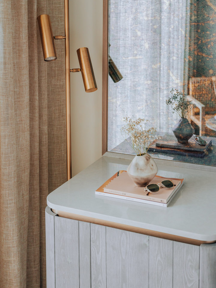 Close up of grey mini fridge with magazine, sunglasses, and flowers on top with mirror behind it and brass standing lamp in background