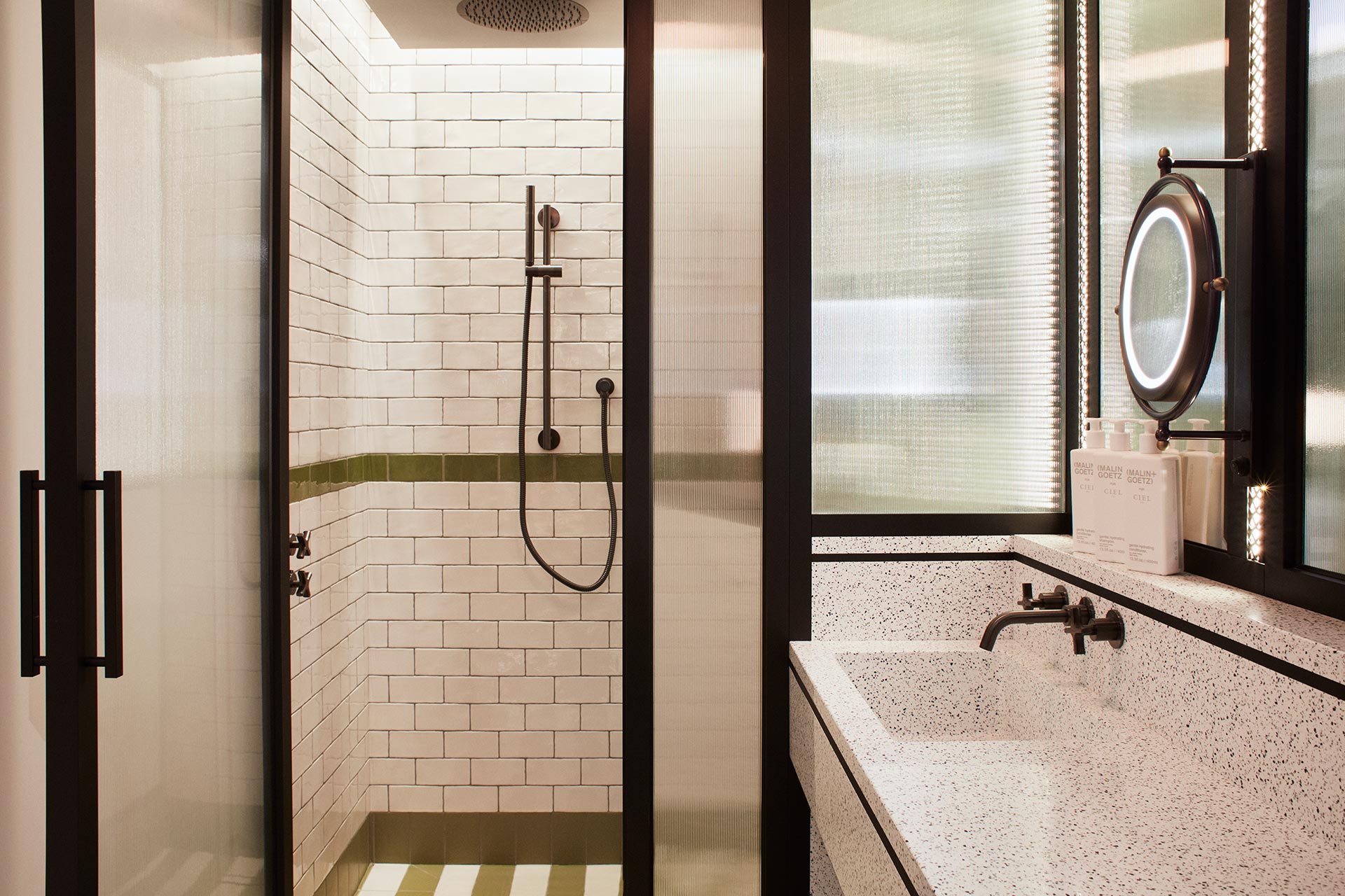 Bathroom with white tiles and standing shower with black lined doors, big white sink, and small circular mirror