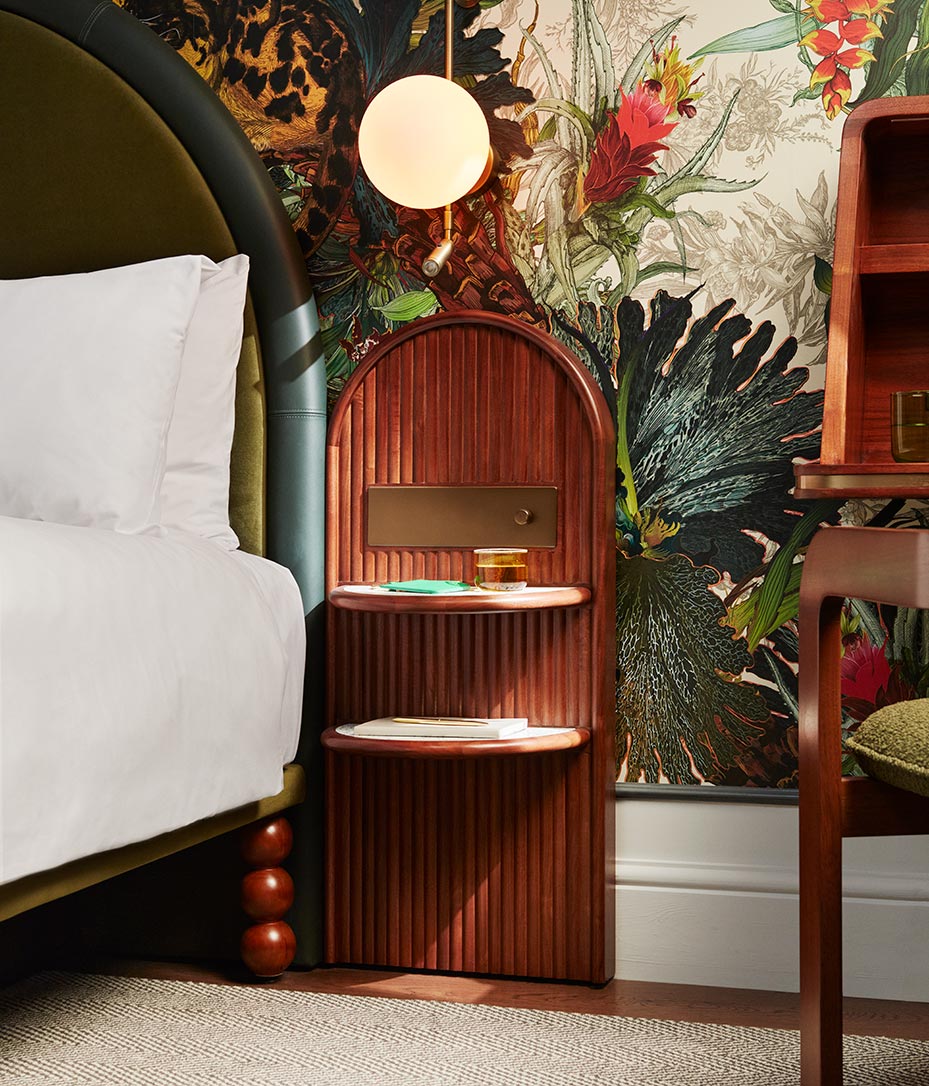 Wooden bedside table with round lighting fixture next to a white bed with green headboard and corner of a wooden desk and chair in the corner
