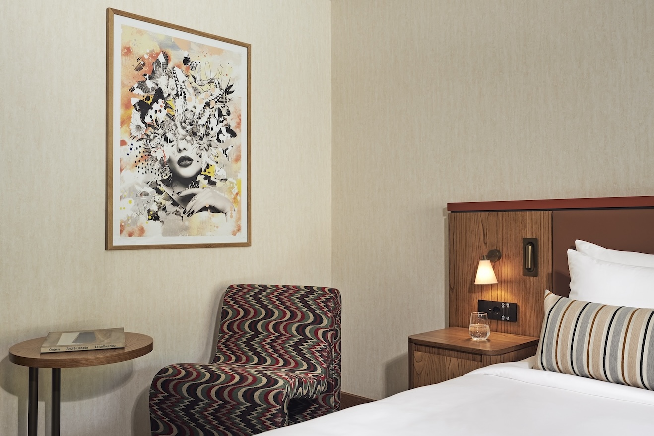 Corner of bed with brown headboard and patterned pillow with zig-zag patterned chair and small table in the corner with framed art on the wall