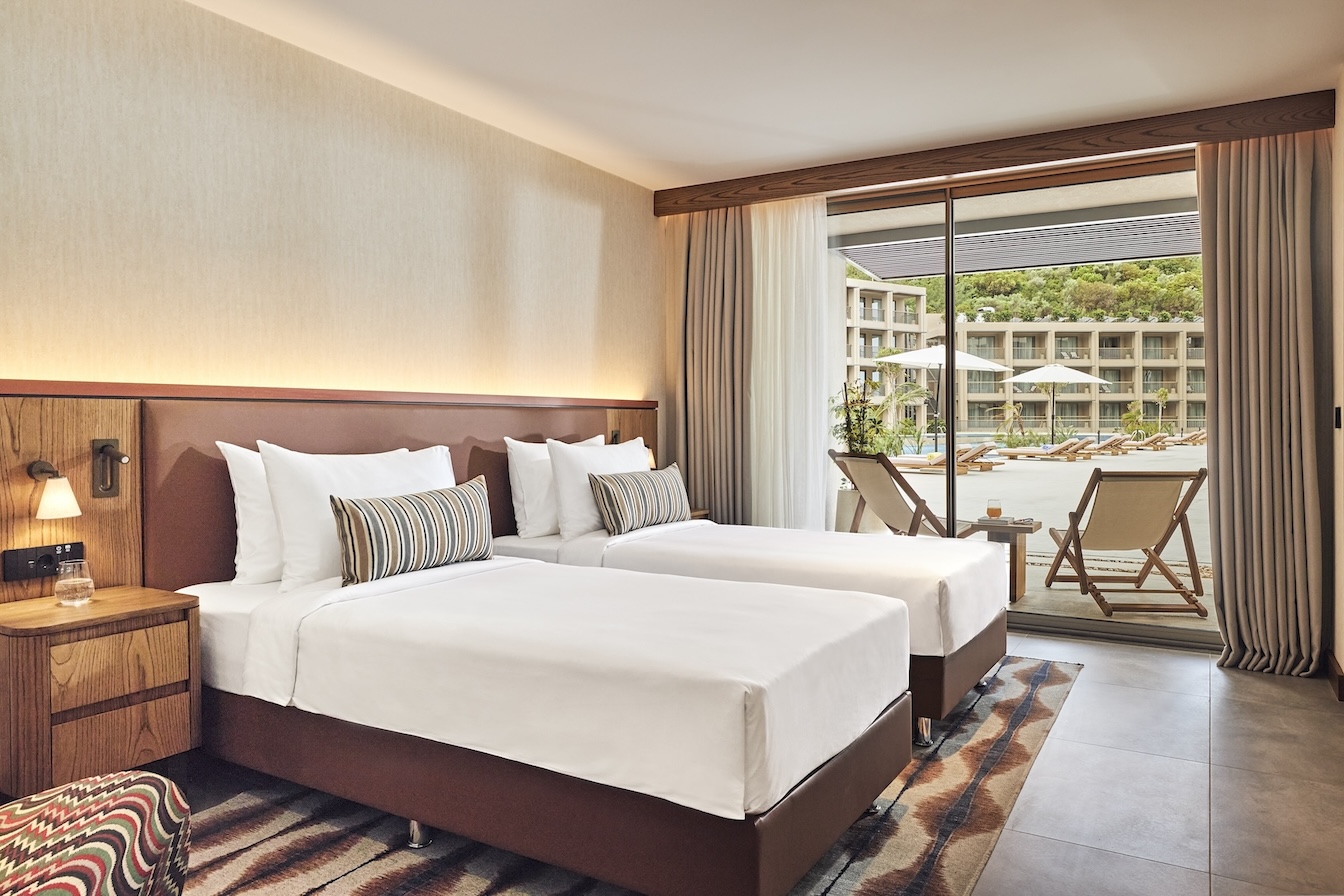 Twin beds with burgundy headboards on a patterned rug with sliding glass doors looking out to a balcony