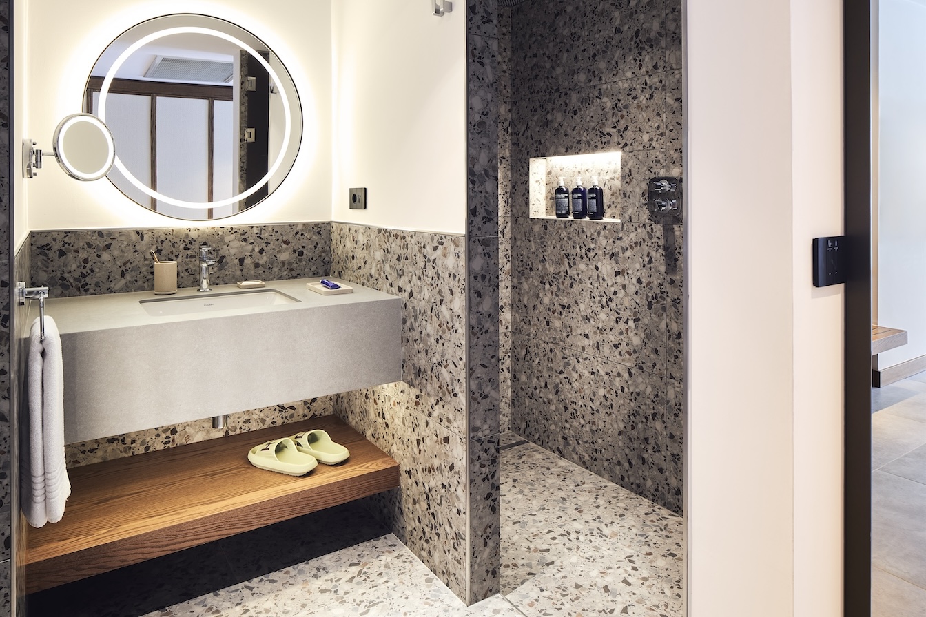Angled view of grey sink and circular mirror with grey marble walk-in shower in the corner