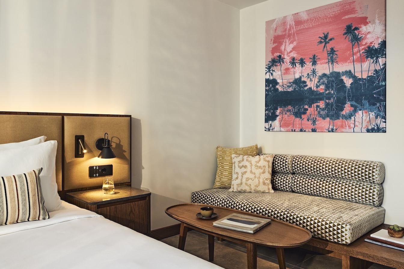 Corner of bed with beige headboard and beige upholstered bench to the right with colorful framed artwork on wall above