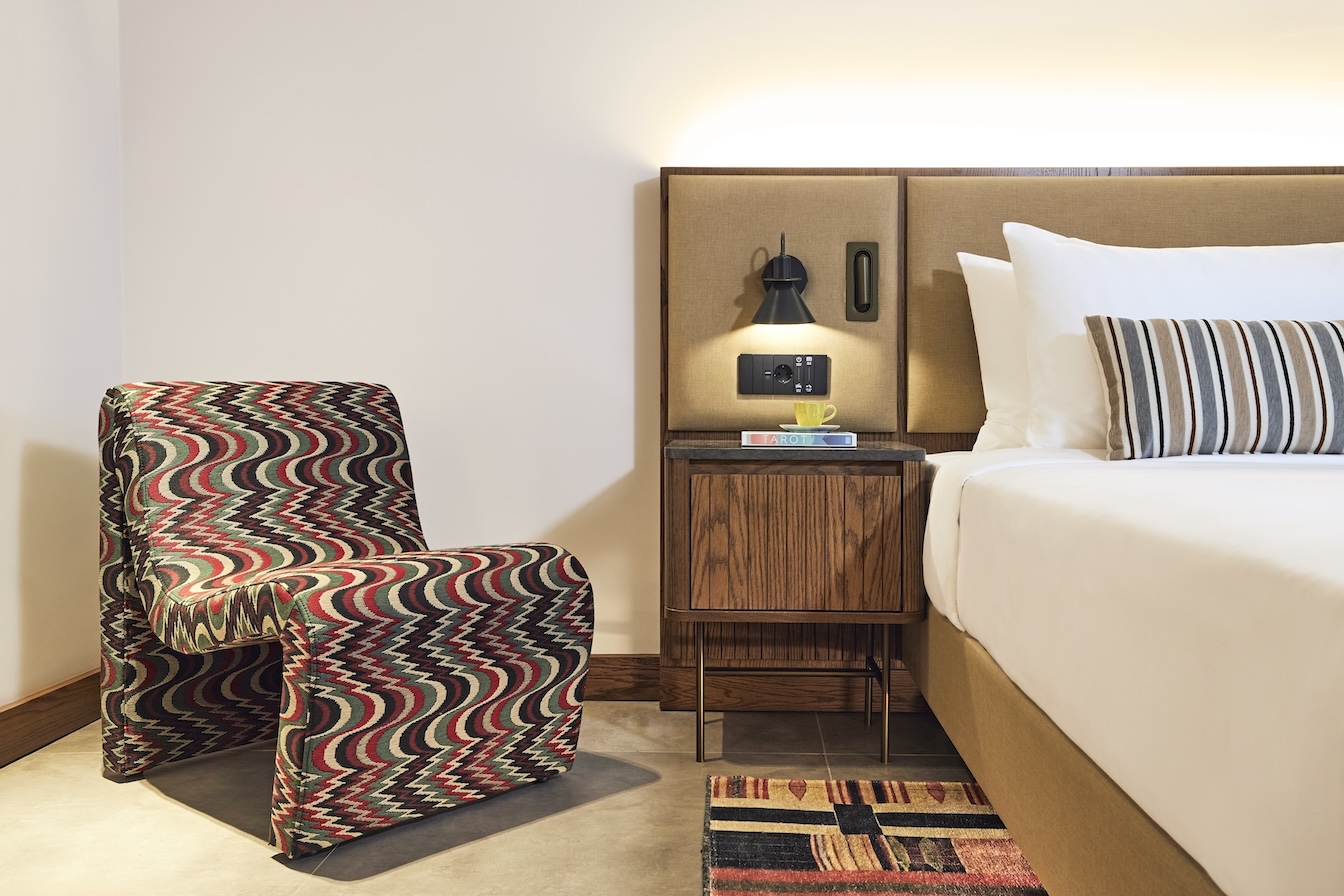 Corner of a bed with beige headboard, side table, small light, and zig-zag patterned chair to the left