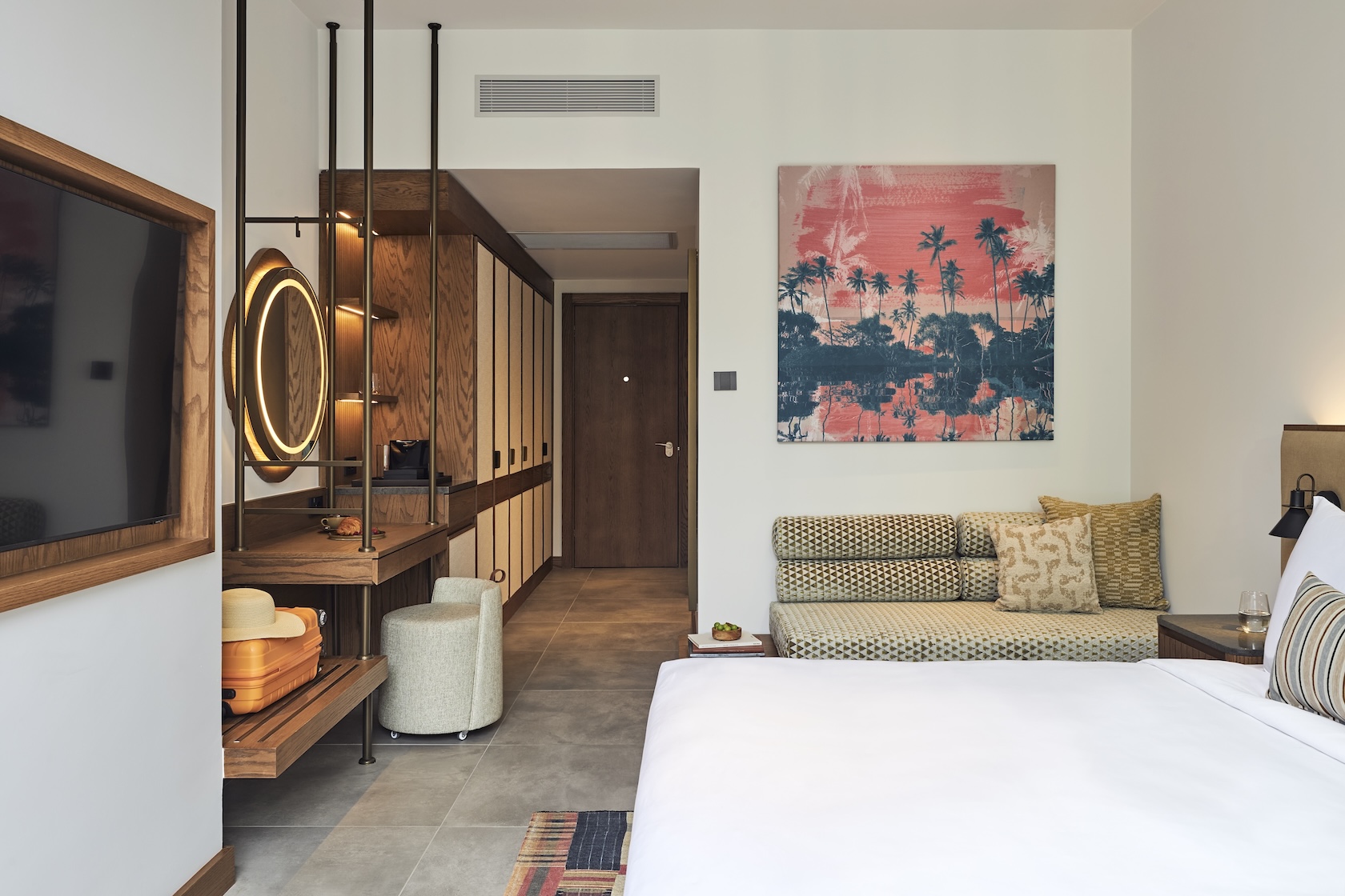 Side view of white bed with plasma screen tv on wall opposite it, an upholstered bench with framed artwork sits above, and a vanity and wardrobe are on the left