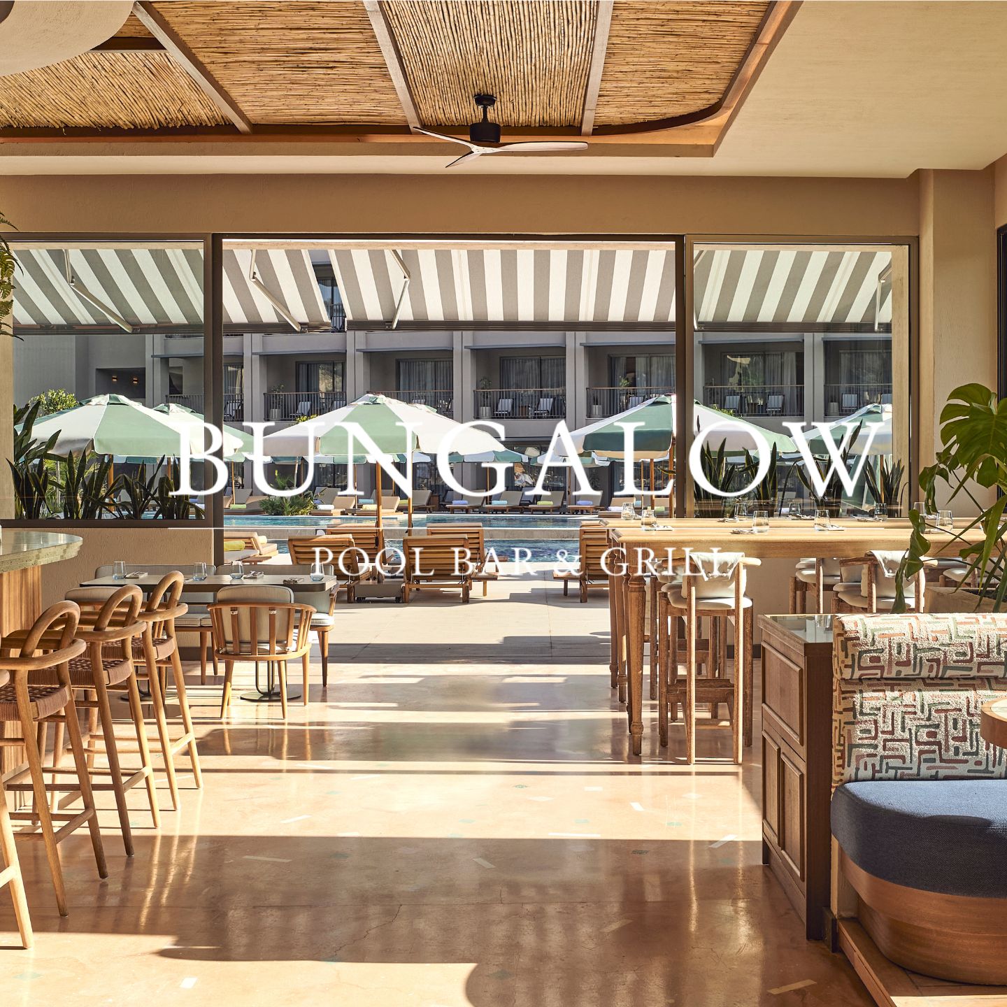 Dining room with booths, wooden chairs, and wooden tables looks out to an outdoor pool bar with striped umbrellas and wooden chaise lounges