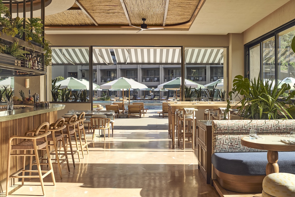 Large dining room with natural sunlight, wooden furniture, cushioned booths, and large door open out to a pool with cabanas