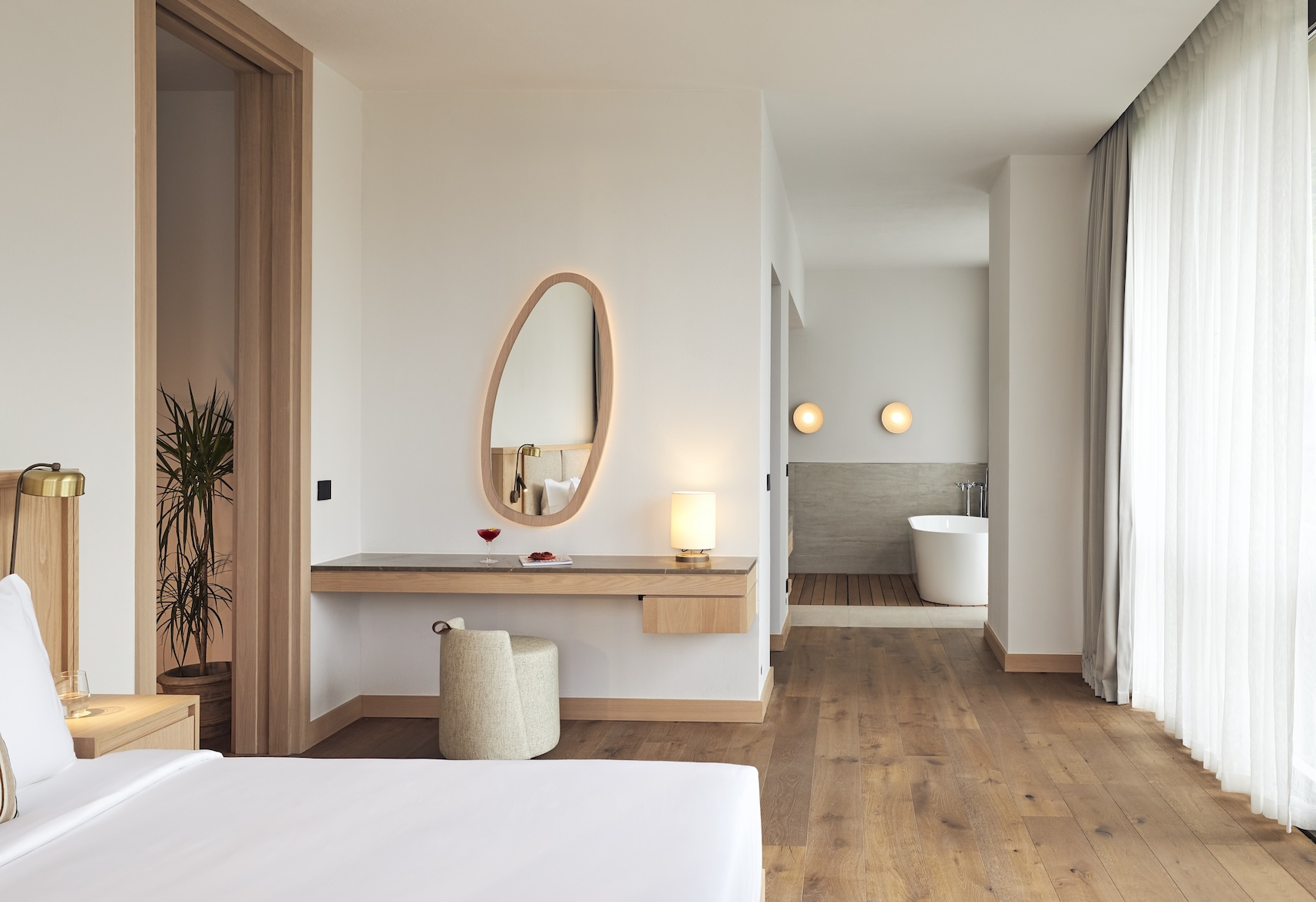 Corner of a white bed with wooden vanity and cream stool and oval shaped mirror on wall