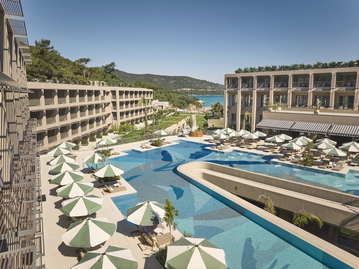 Overhead shot of a pool with interesting design at the bottom and white and green umbrellas over lounge chairs with a forest and sea in the far distance