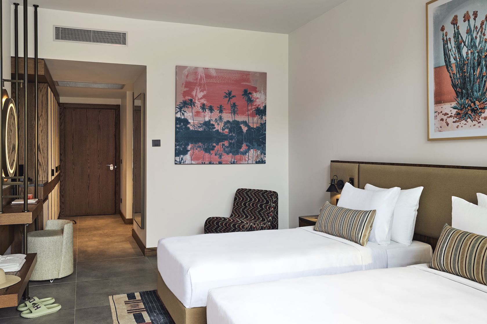 Two twin beds with beige headboard with chair in the background and framed artwork on the wall