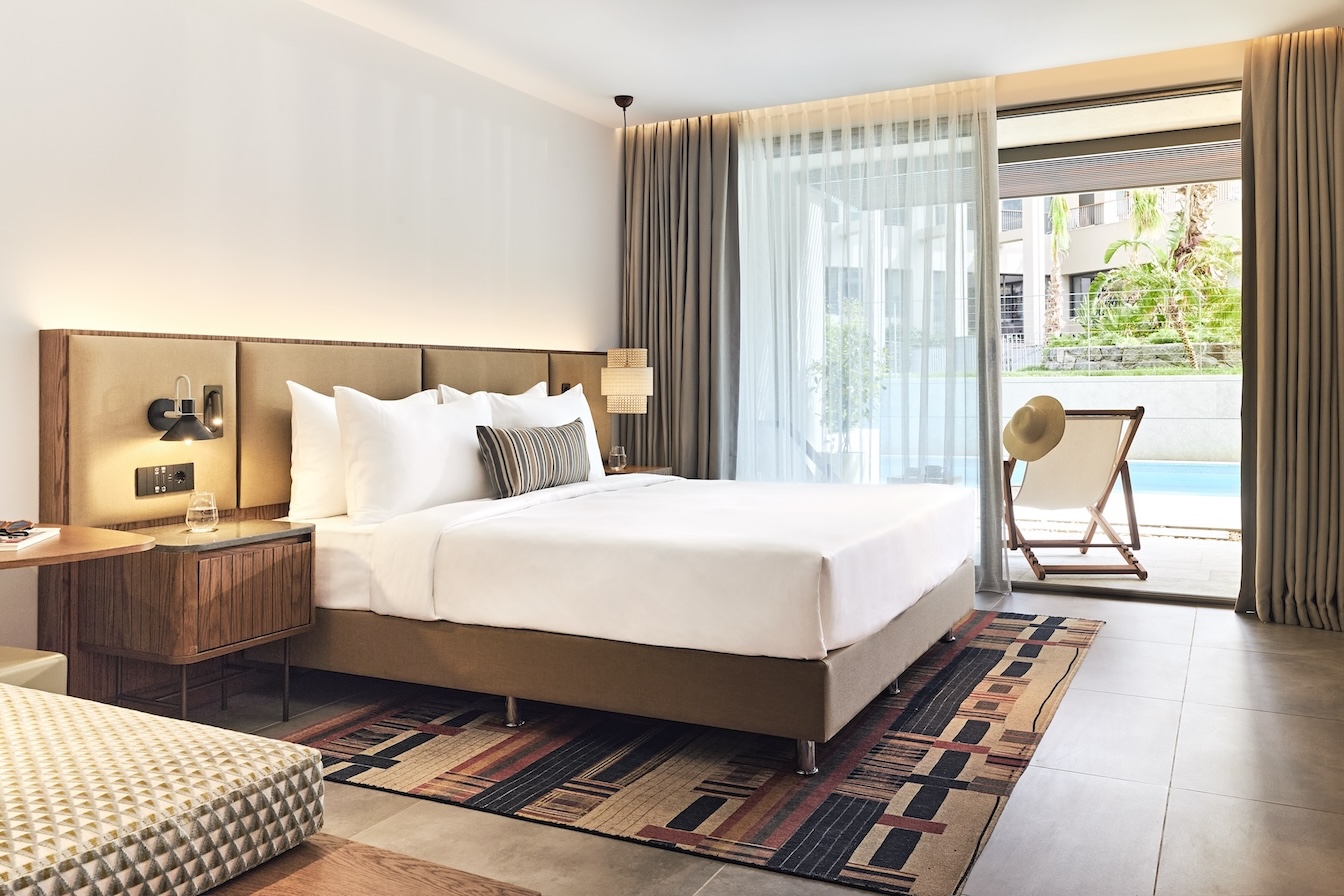 Large white bed with beige headboard on patterned rug with open glass door looking out to balcony with chair