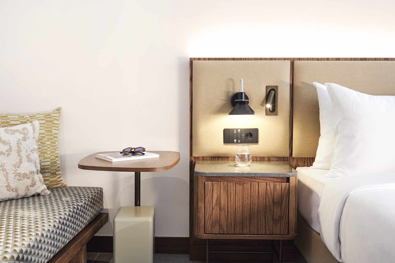 Close up of white bed with beige headboard, small bedside table, small lamp, and upholstered bench to the left