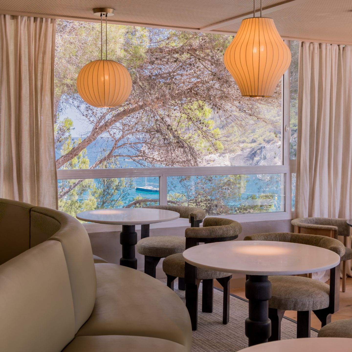 Dining room with grey booths, small white round tables, hanging lantern lights, and a large window looking out to the sea
