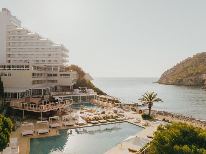Two beautiful pools with cabanas and palm trees with a white building and a sea with mountains in the background