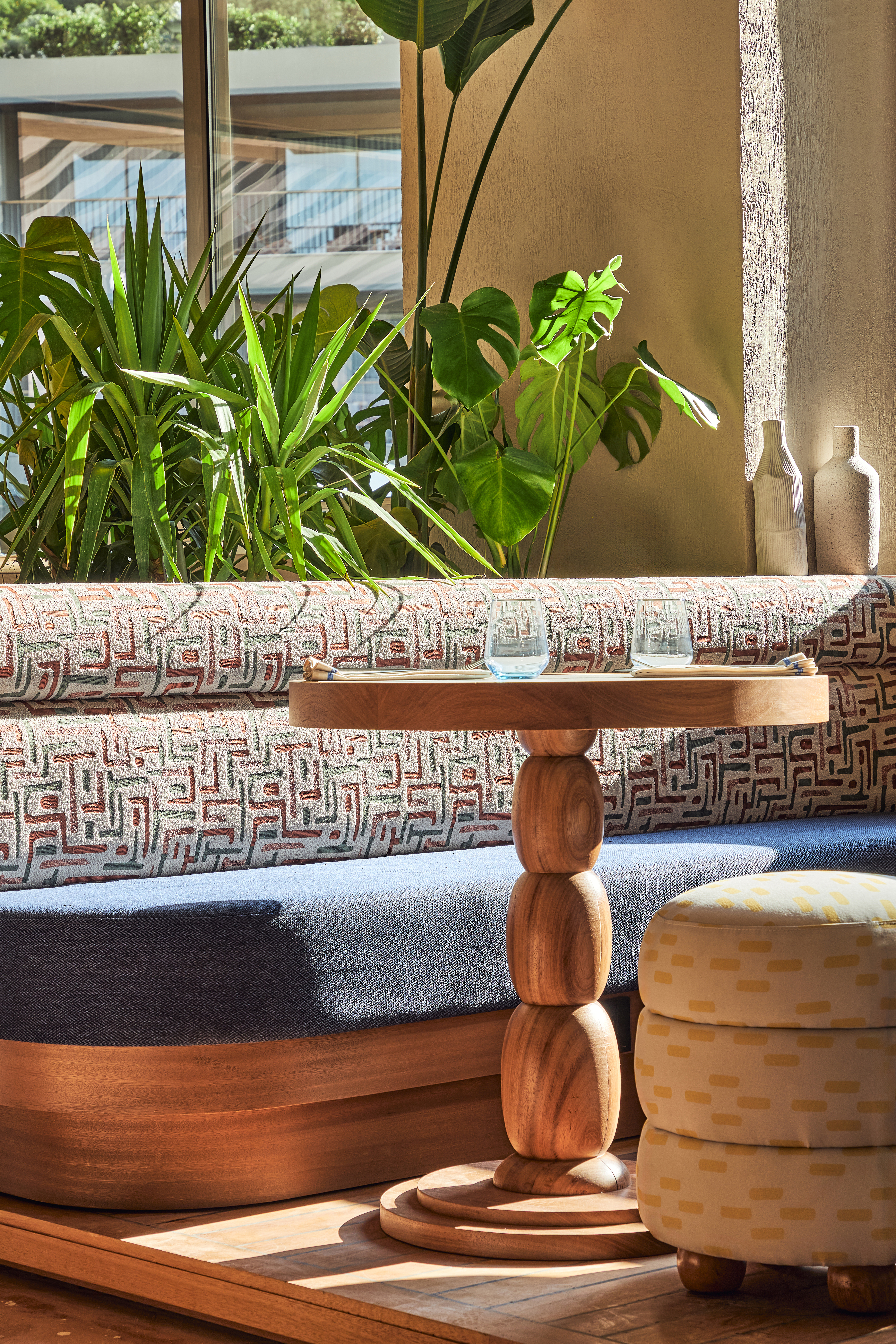 A hotel lobby corner with a wooden table, sofa, a stool and some greenery