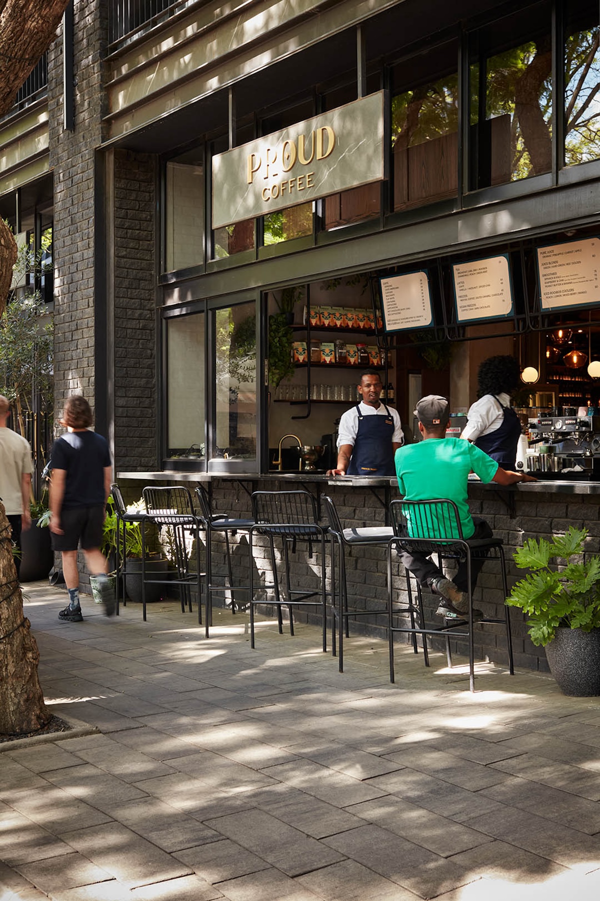 The barista is taking an order at the Proud Coffee, with a customer sitting in one of the bar stools.