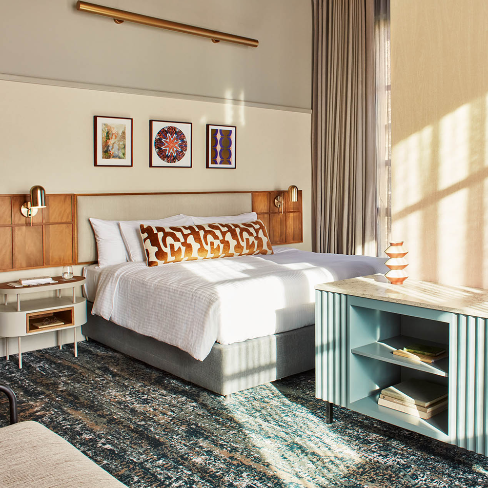A king-sized bedroom, with art pieces hanging on top of the bed, a pillar to the right of the image, with a blue designed table that has shelves for additional storage.