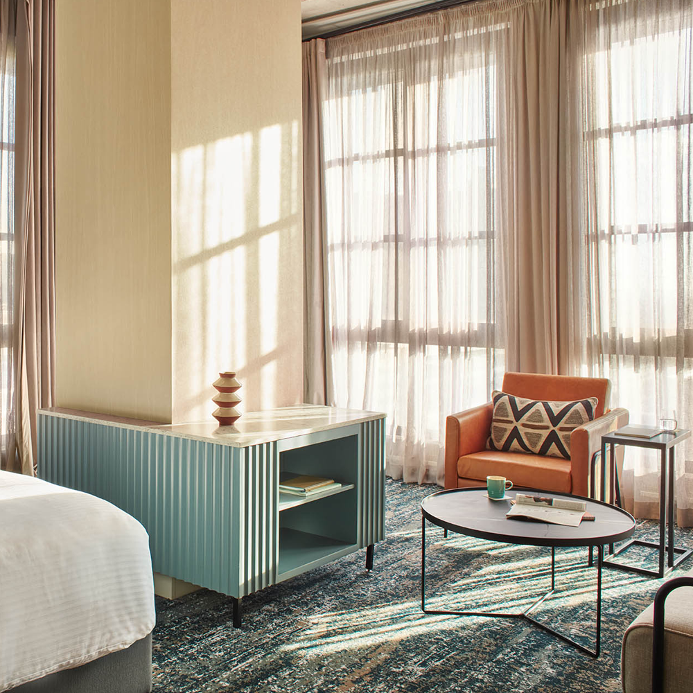 Sunlight pouring through the sheer curtains, the 1-seater leather brown couch, a side table, and a round, black table in front of the couch. There is also a blue designed table with shelves for additional storage.