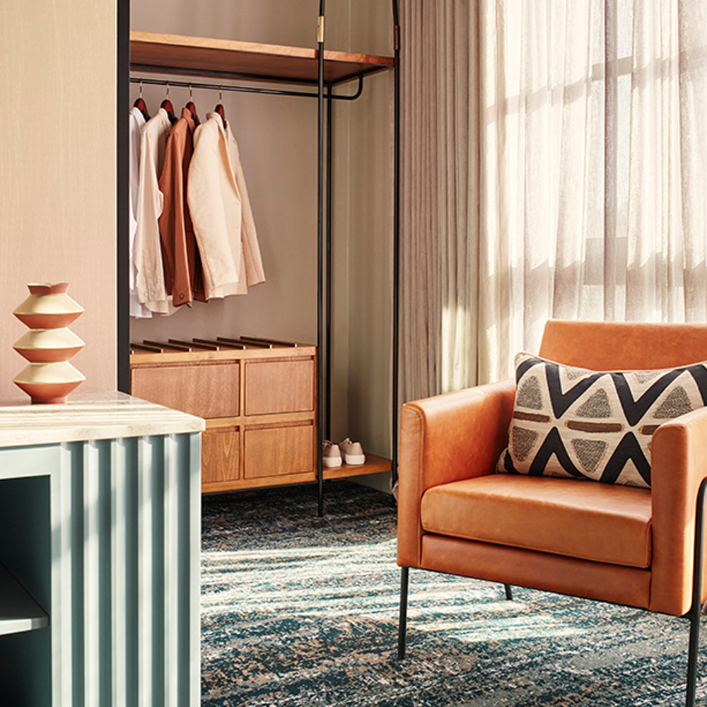 Sunlight pouring through the sheer curtains, a 1-seater brown, leather couch and an open wardrobe with wooden drawers at the bottom in the back of the image.