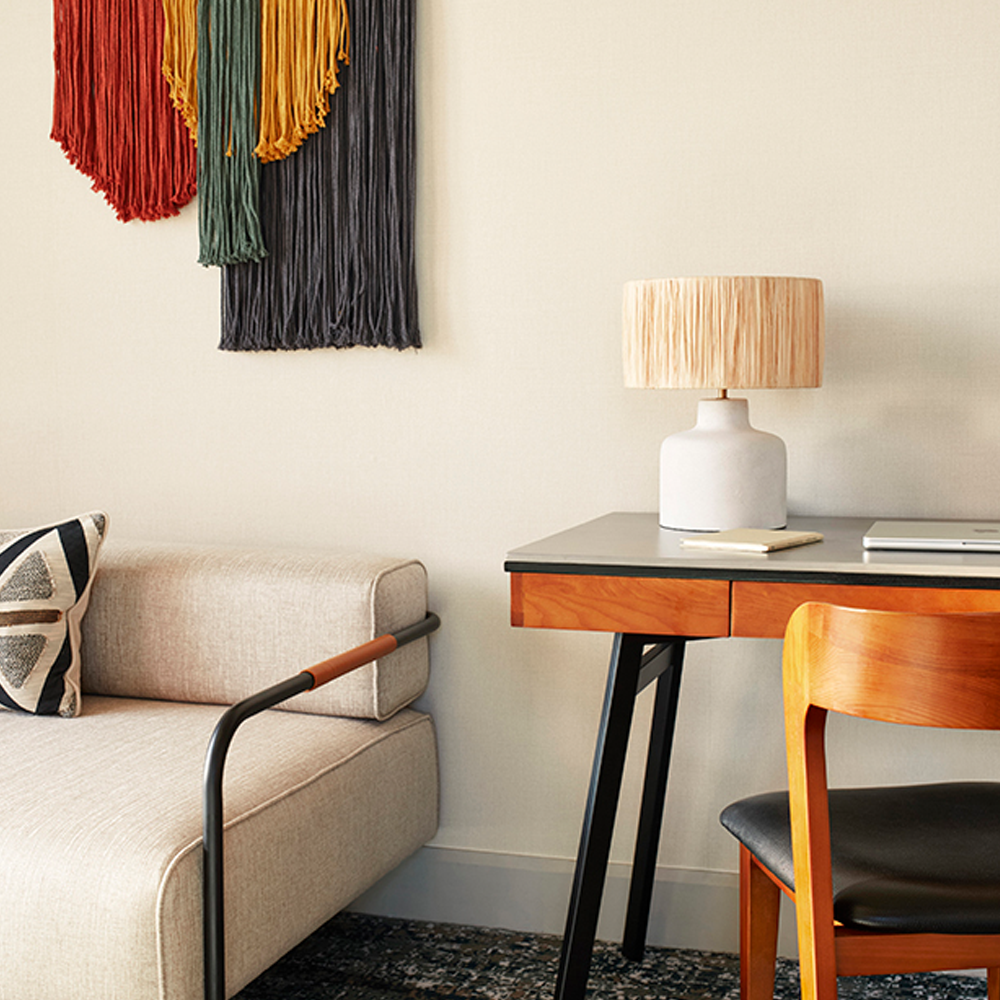 A 3-seater couch, with hanging wall art, and a desk that can be used for work or as a makeup area, and a lampshade on top of the table.