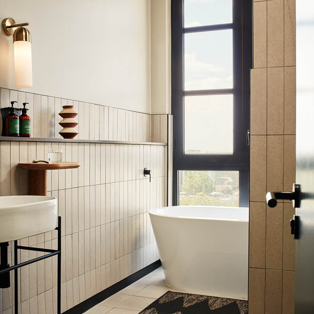 A white sink to the left, with a light next to the mirror, and a bathtub with ample light pouring in through the window behind.